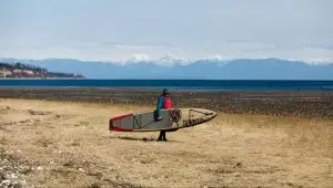 Norm Hann carrying SUP on beach with mountains in the background
