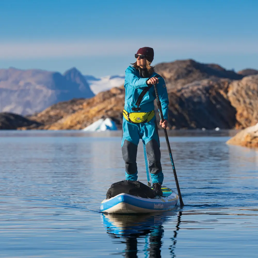 Person standing on a blue-and-white stand-up paddleboard, paddling on calm water with rocky mountains in the background, wearing a blue jacket and yellow waist pack.