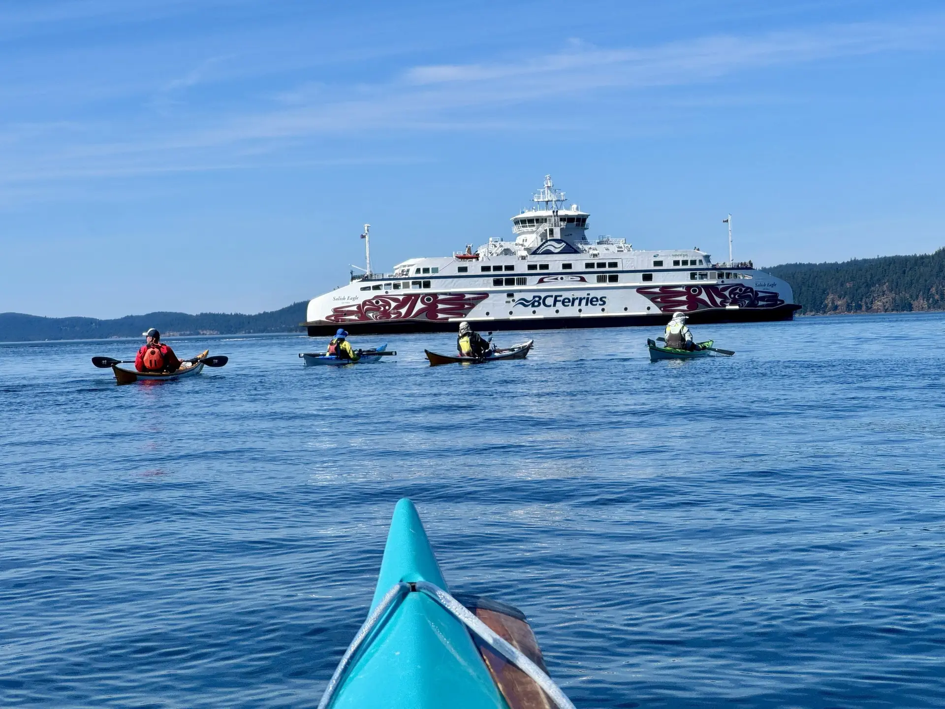 Perspective from a turquoise kayak bow toward several kayakers paddling on calm blue water with a large white BC Ferries vessel in the distance and forested hills beyond.