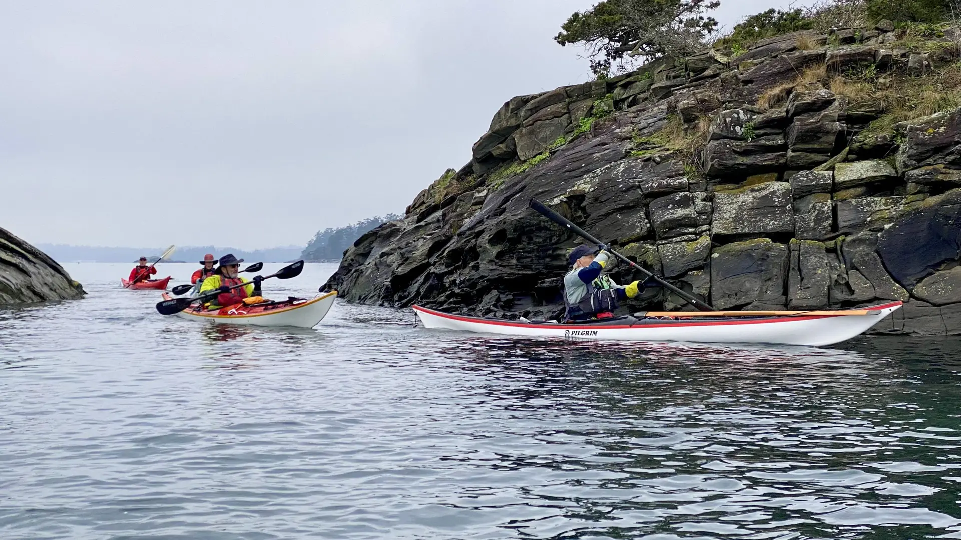 Three kayakers in red life jackets paddle near a rocky, mossy shoreline on calm water under a cloudy sky.