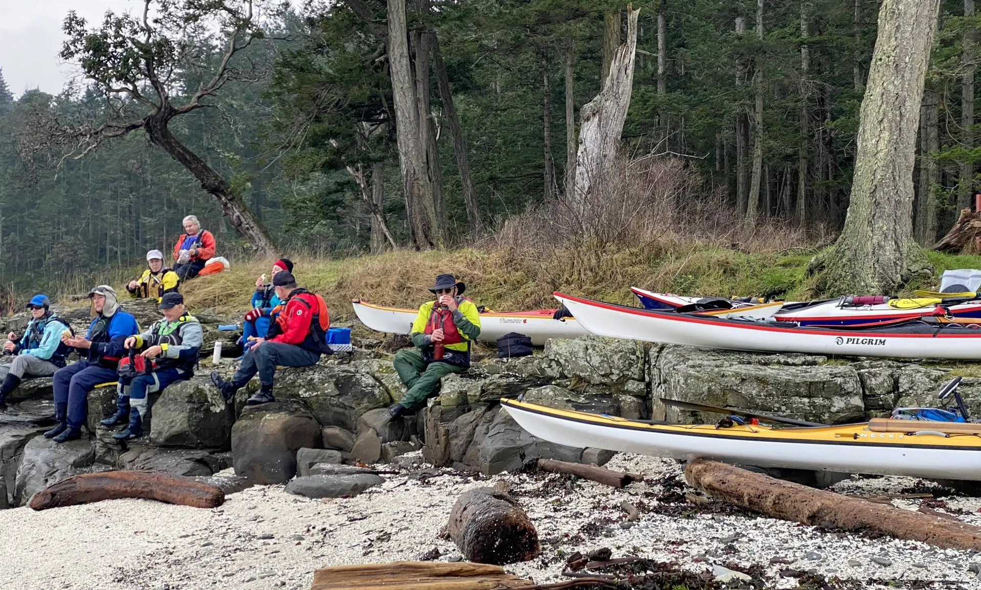 Group of hikers in bright jackets sitting on a rocky shore with colorful kayaks lined up along the water's edge.