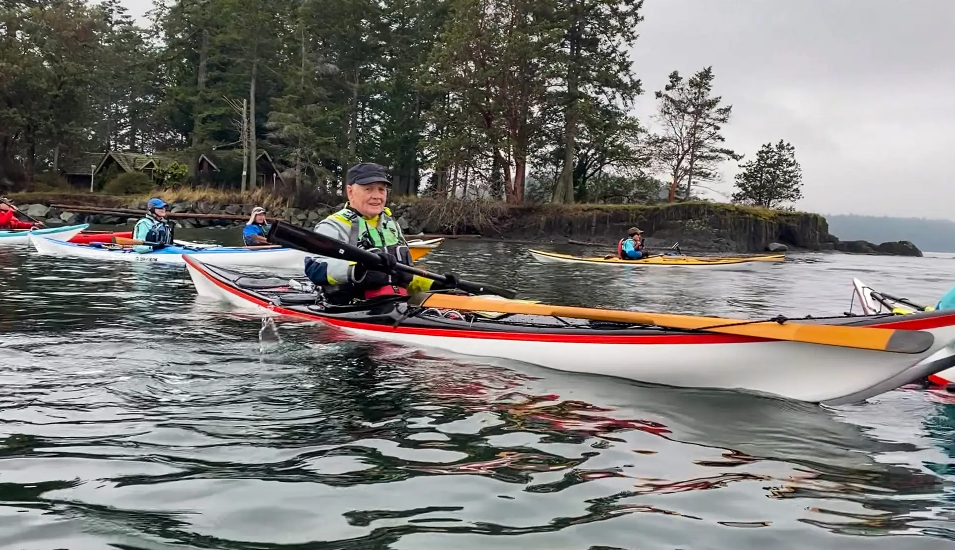 Group of kayakers paddling near a rocky shoreline on a calm day; foreground kayaker wearing a green life vest and cap.