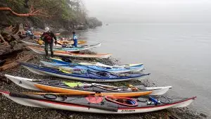 Colorful kayaks lined up on a pebbled rocky shore, with people preparing gear near the calm, misty water.
