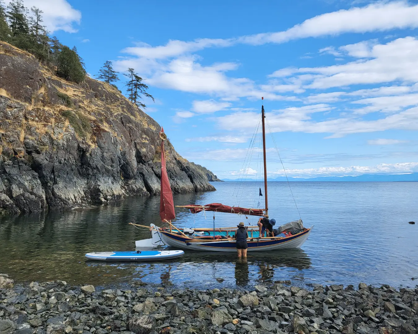 Sailboat moored near a rocky shoreline with two people on board and a paddleboard floating nearby in calm blue water under a clear sky