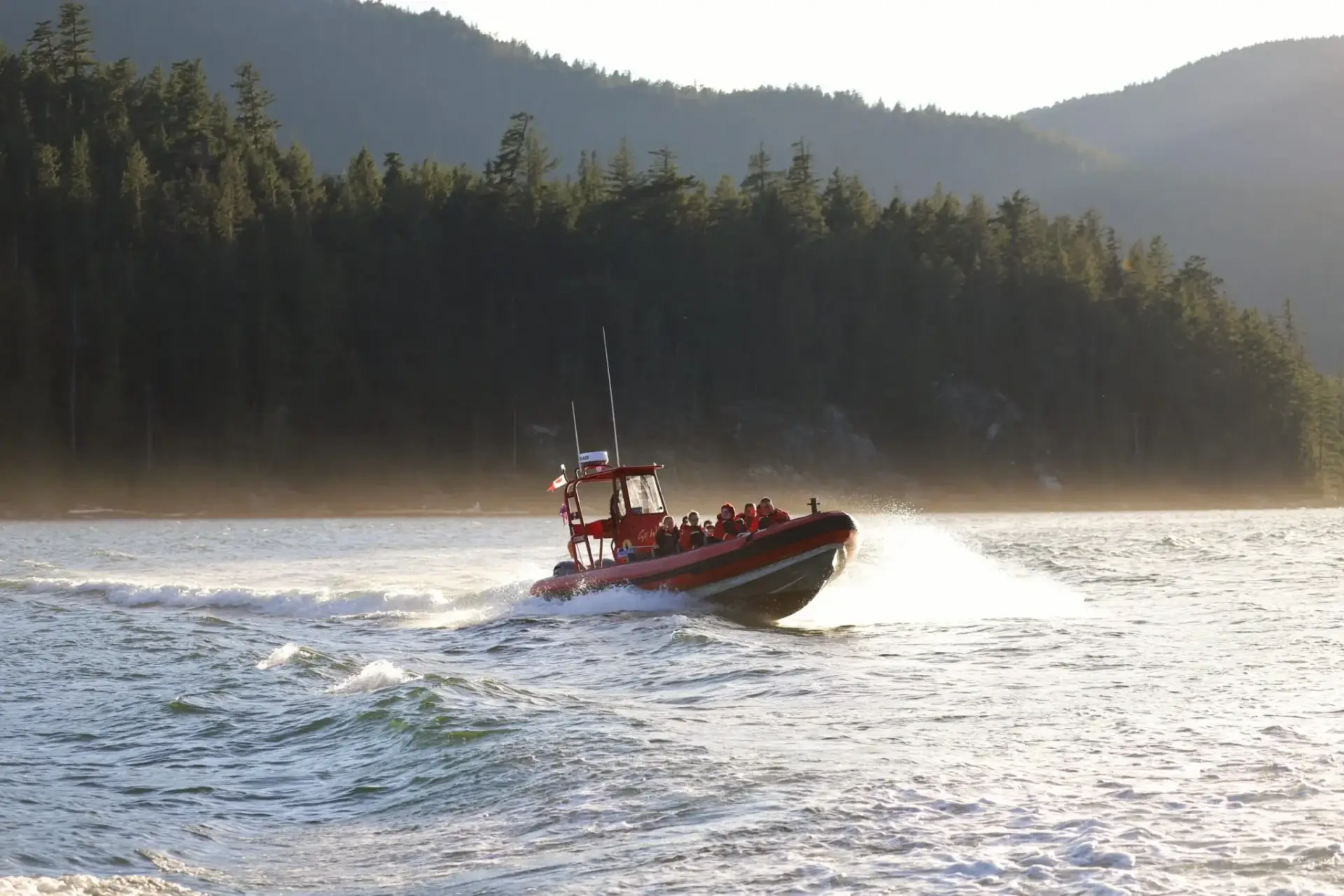 Red inflatable rescue boat speeding across a lake with passengers in life jackets, forested hills in the background.