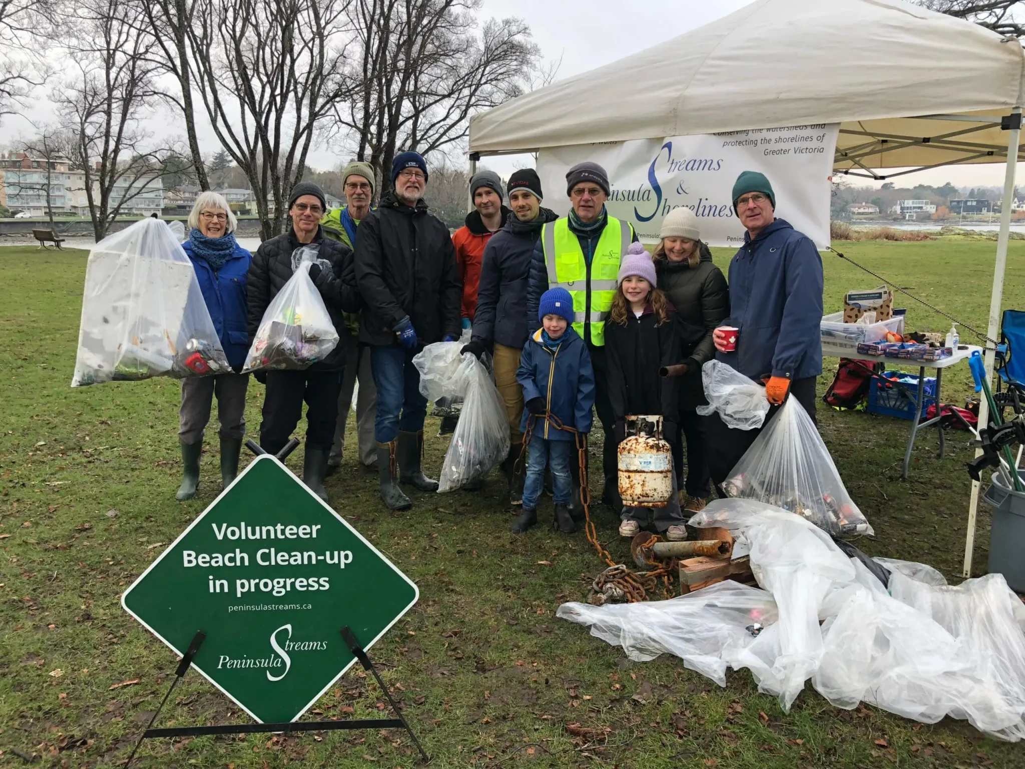Group of volunteers posing together outdoors at a beach cleanup event; several hold large clear bags of trash, with a green “Volunteer Beach Clean-up in progress” sign in the foreground and a tent behind them by a grassy area near water.