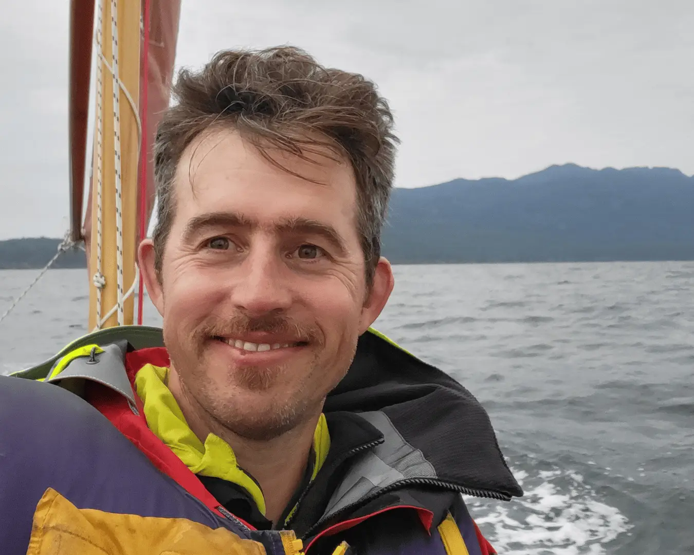 Smiling man wearing a yellow and red life jacket on a boat, gray sea with distant mountains in the background.