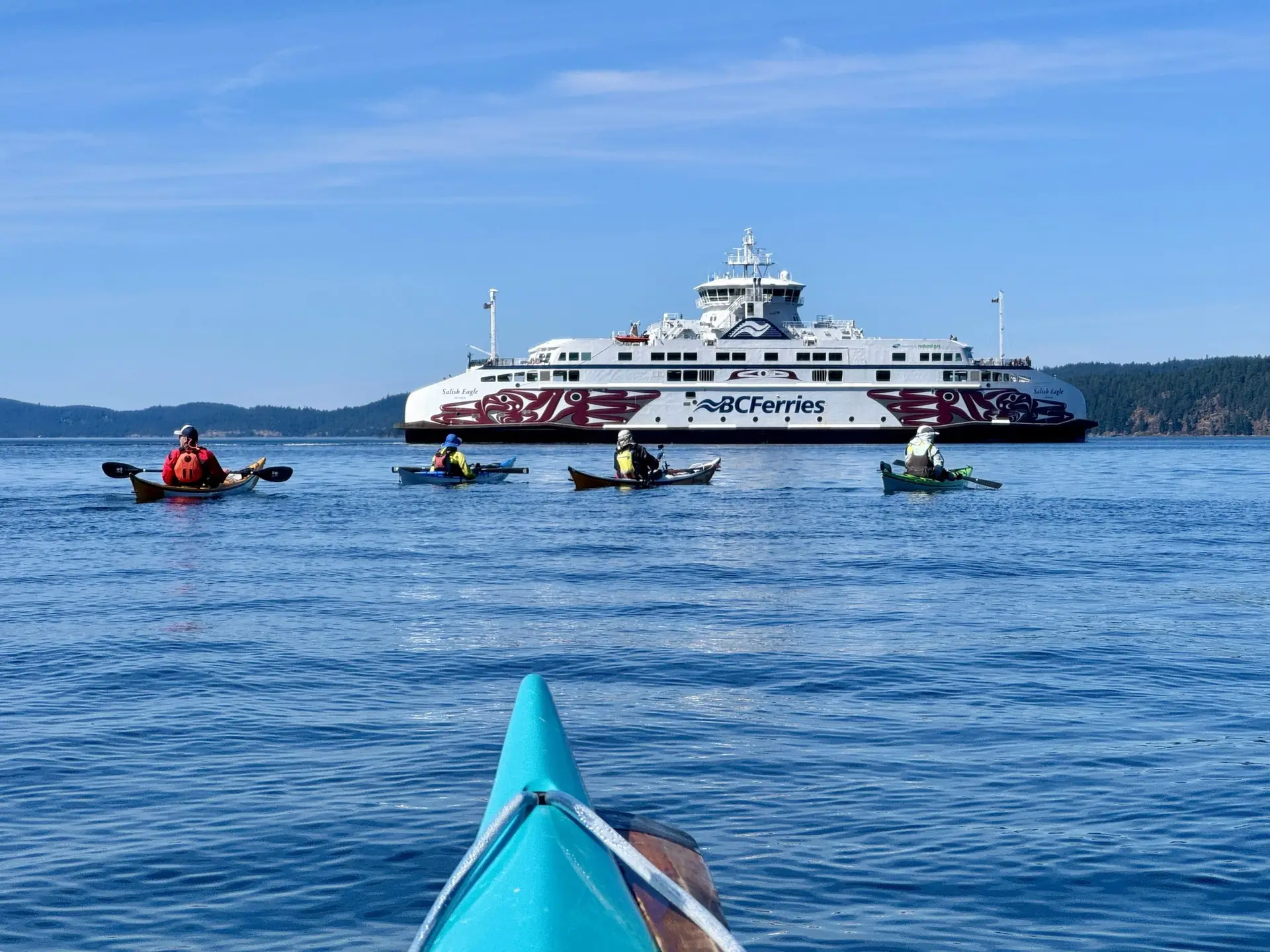 Perspective from a turquoise kayak paddling toward a large BC Ferries ship on calm blue water, with several kayakers nearby and a forested shoreline in the distance.