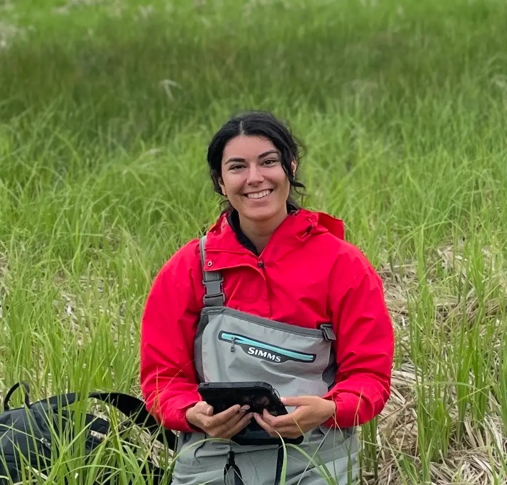 Woman in a red jacket and gray chest waders sits in tall grass, holding a tablet with a smile.
