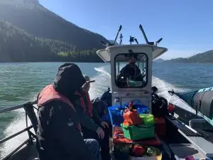 Two people in a small motorboat wearing life jackets, surrounded by colorful gear on a calm lake with forested mountains in the background.