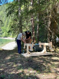 Two men assemble a wooden frame on a small platform beside a lake, surrounded by tall trees in a forested area