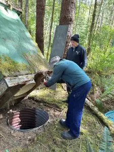 Two men in a forest work near a mossy, curved green hull or barrel leaning against a tree.