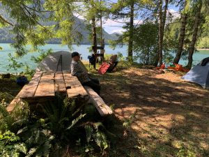 Campsite by a turquoise lake with a grey tent, a man sitting on a wooden platform, and camping gear among trees.