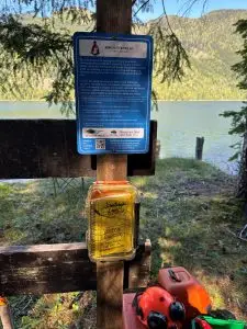 Blue informational sign mounted on a wooden post by a lake, with forested hills in the background.