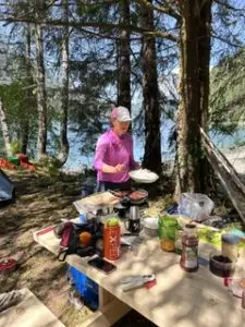 Woman in a pink shirt and cap cooking at a campsite table with condiments and a grill beside a wooded lake.