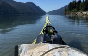 View from a kayak looking toward distant mountains across a calm lake, with a map laid on the deck and paddles visible in the foreground.