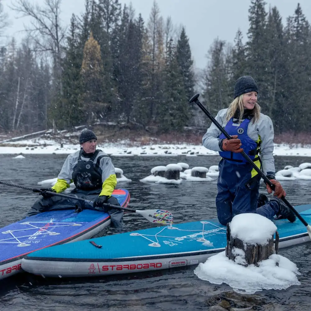 Two people on stand-up paddleboards in a snowy lake, wearing life jackets and cold-weather gear.