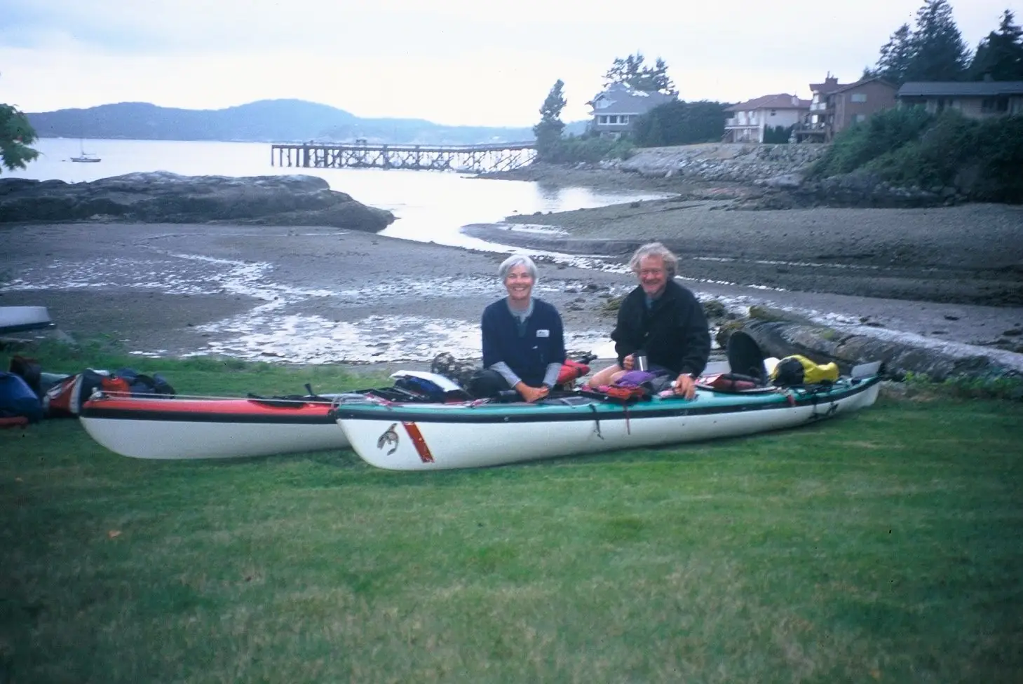 Two people sit in a white and teal tandem kayak on a grassy shoreline with gear laid out and a pier in the distance.