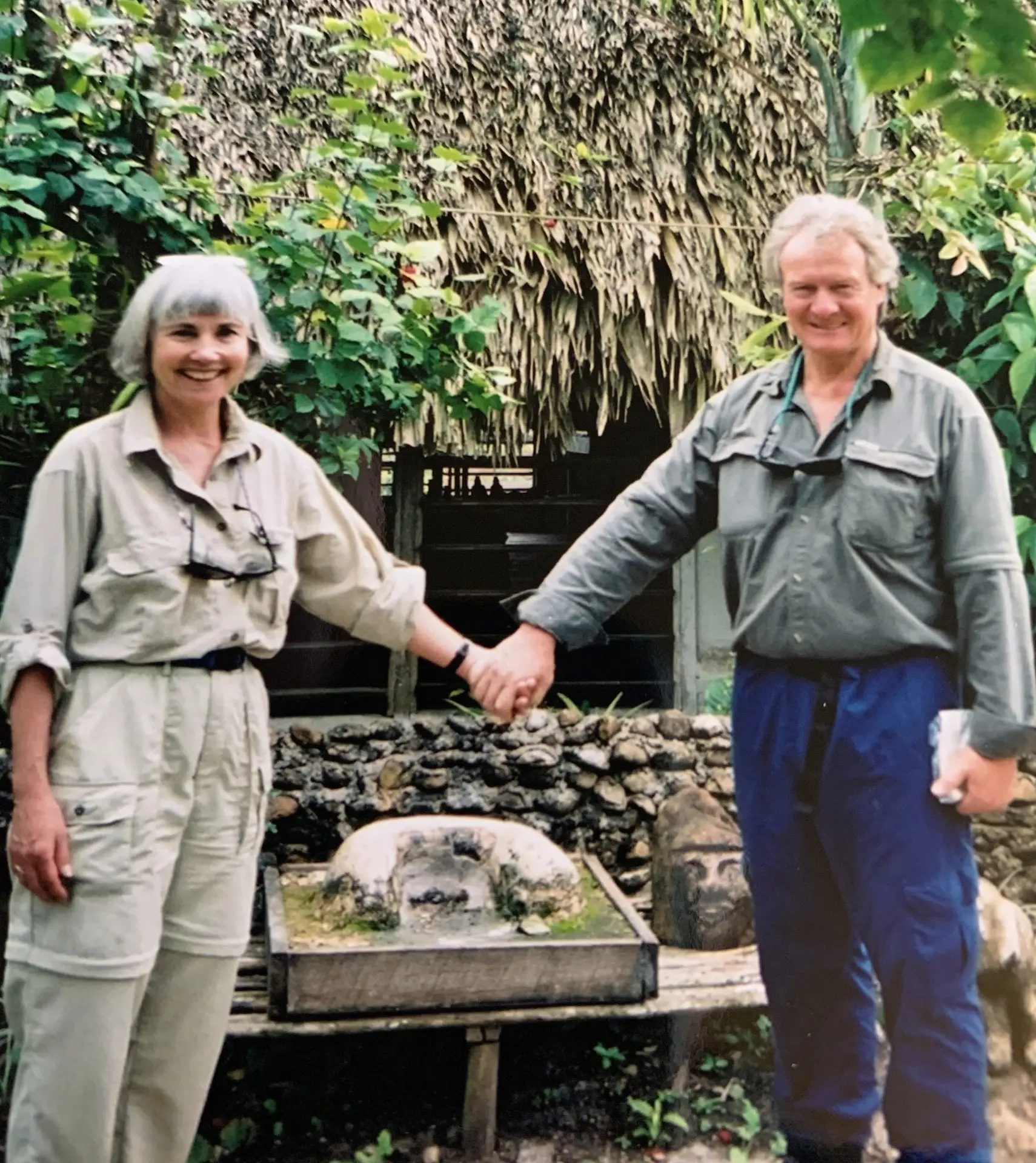 Two adults in khaki uniforms stand outdoors, holding hands and smiling with a leafy garden backdrop behind a stone feature.