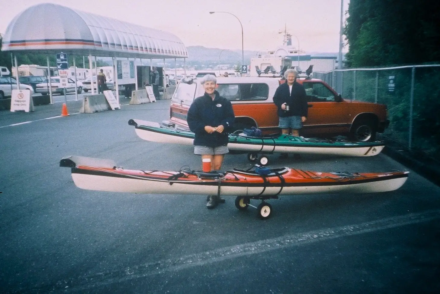 Two people stand beside a red and a white canoe/kayak trailer in a parking lot by a waterfront facility, each with a boat gear setup nearby.