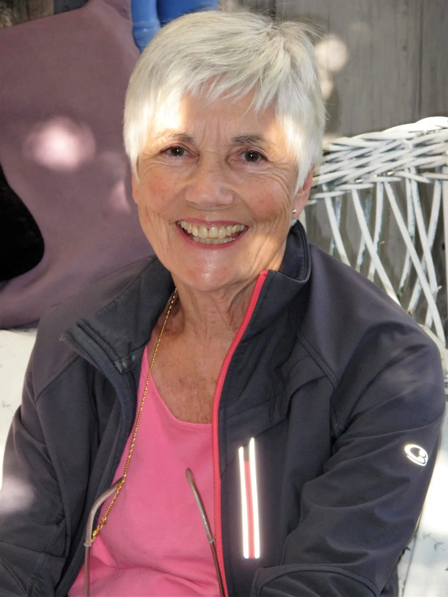 Smiling older woman with short white hair wearing a pink top and dark jacket, seated outdoors.