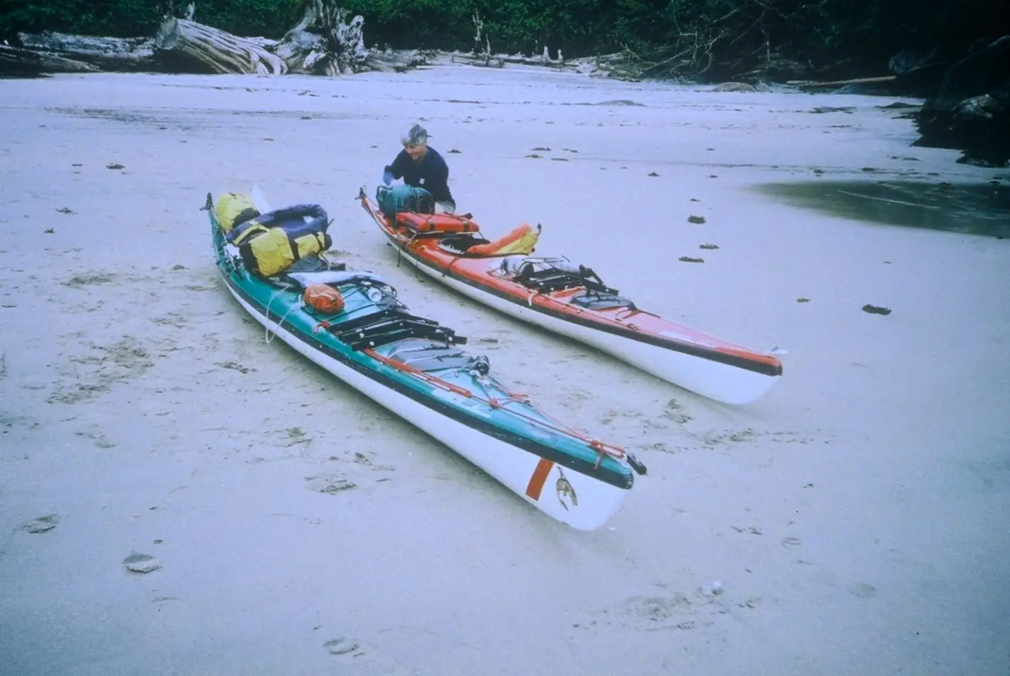 Two colorful kayaks with gear sit on a sunny, sandy beach; a person works on equipment beside them.