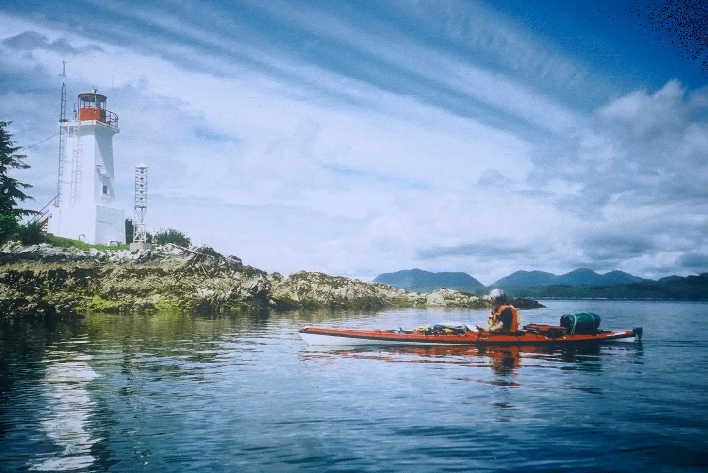 White lighthouse on rocky shore with a person in a red kayak gliding on calm water, mountains in the distance under a blue sky