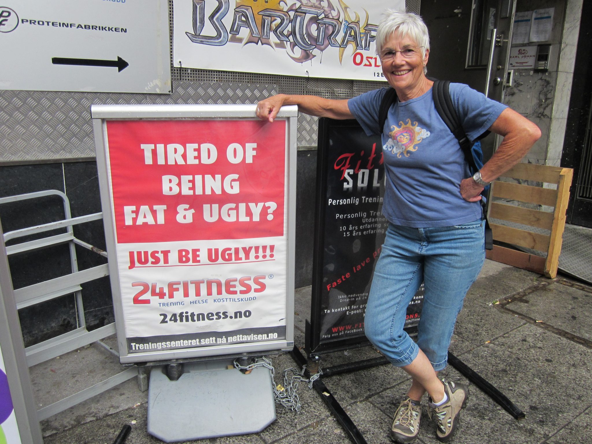 Smiling woman with short white hair and a backpack stands beside a bold red sign that reads: 'TIRED OF BEING FAT & UGLY? JUST BE UGLY!!!'