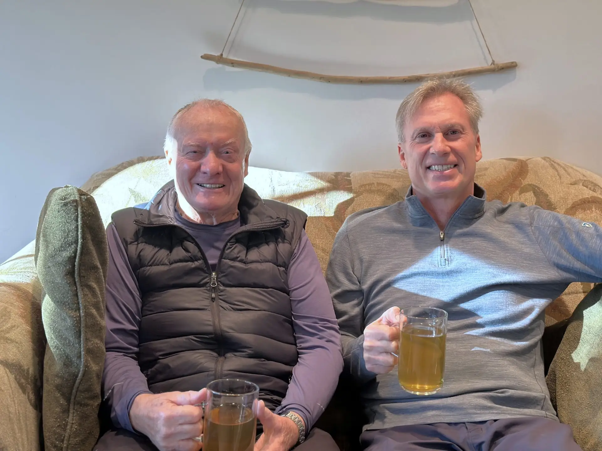 Two smiling men sit on a patterned sofa indoors, each holding a tall glass of beer toward the camera.