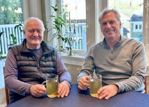 Two older men smiling at a table, each holding a glass of pale green beverage indoors near a window with star garlands in the background.