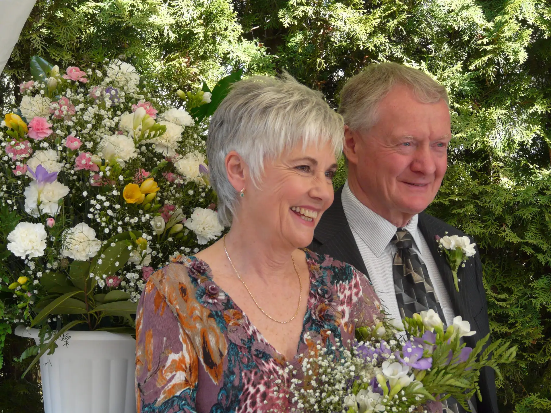 Smiling elderly couple in formal attire posing among a lush floral backdrop at a wedding event.