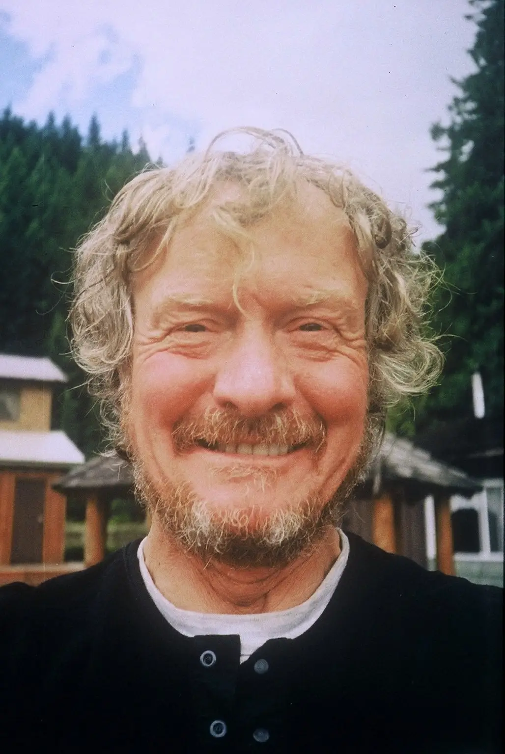 Close-up of a smiling man with gray hair and beard outdoors, with trees and wooden buildings in the background.