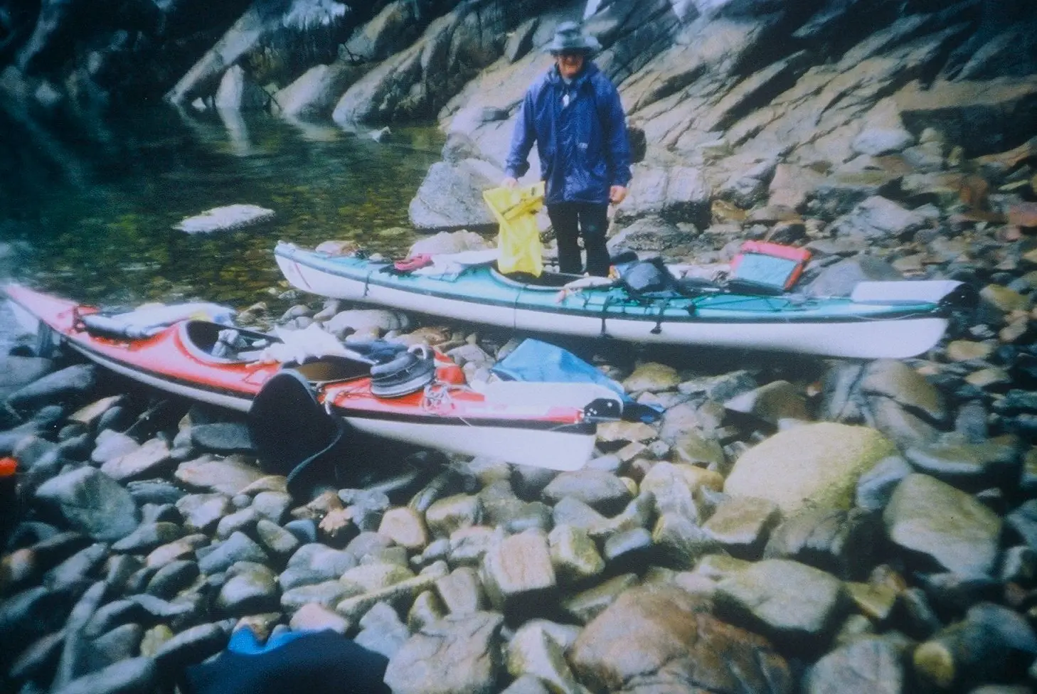 Person in a blue rain jacket standing in a kayak on a rocky shoreline with gear and backpacks nearby.